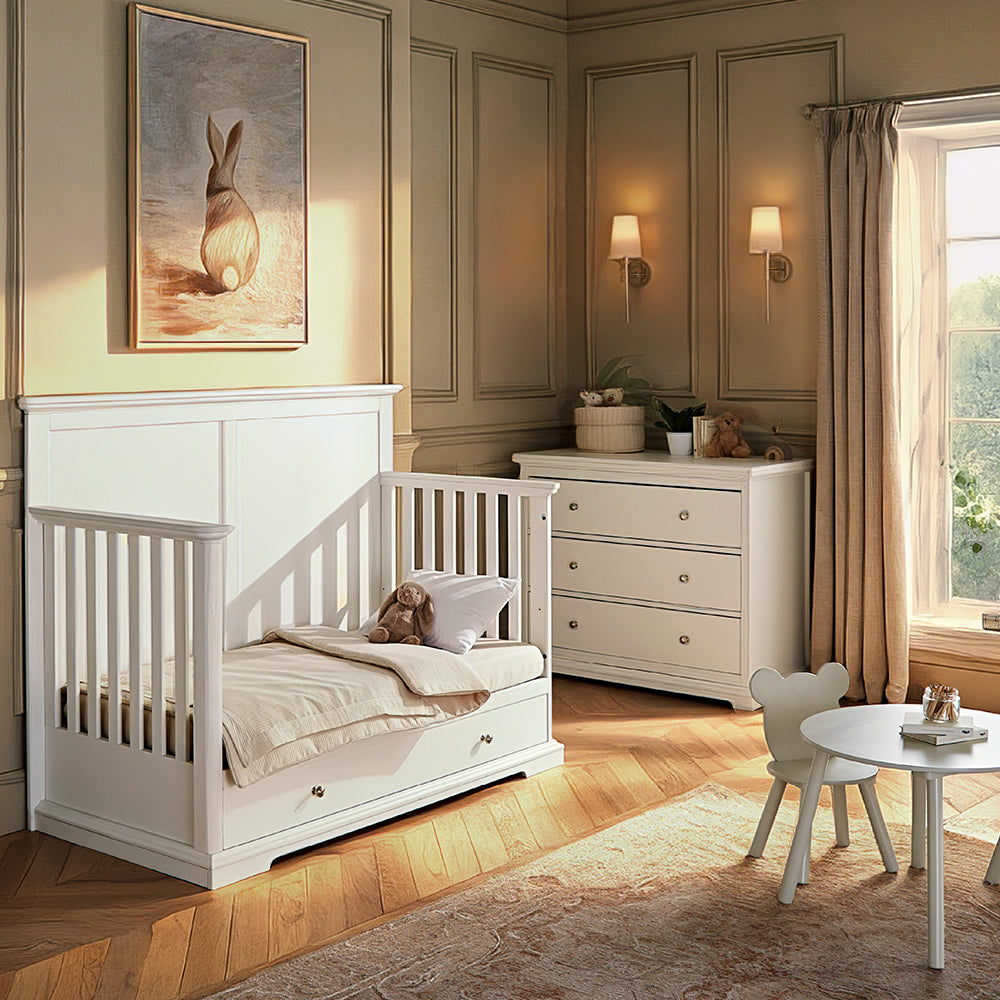 A white wooden toddler bed with a guard rail and built-in storage drawers, styled in a warm, classic children's bedroom with beige walls, a matching dresser, and a small white chair.
