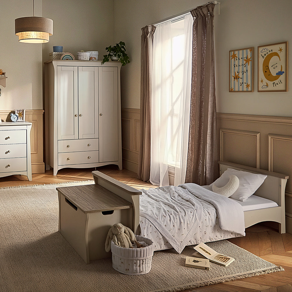 A styled children's bedroom featuring a light wood single bed with white bedding, a matching wooden storage bench, a cream-colored double dresser, and a tall double-door wardrobe. The room has warm beige walls with wainscoting, brown curtains, and a textured beige rug on a wooden floor.