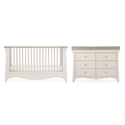 A baby nursery furniture set featuring a convertible crib and a six-drawer dresser in a warm off-white finish with light natural wood tops and small gold knobs, shown against a plain white background.