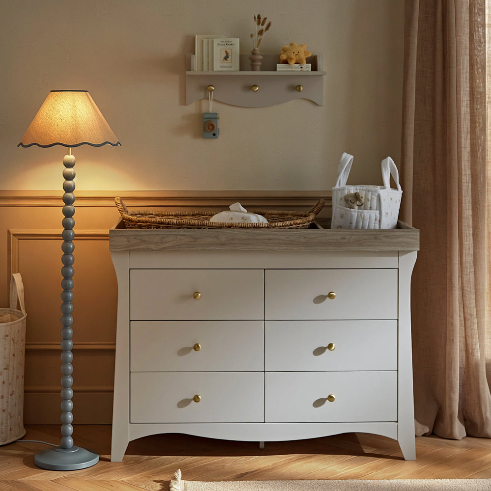 A white dresser with six drawers and gold knobs, styled in a nursery or children's room next to a blue beaded floor lamp, with a changing tray on top.