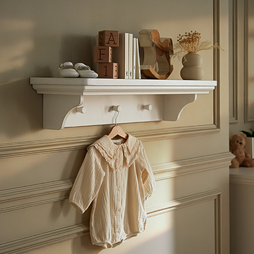 A cream-colored wall-mounted shelf with decorative brackets and two hooks, styled with baby shoes, books, wooden blocks, and dried flowers, with a matching cream baby romper hanging from one hook.