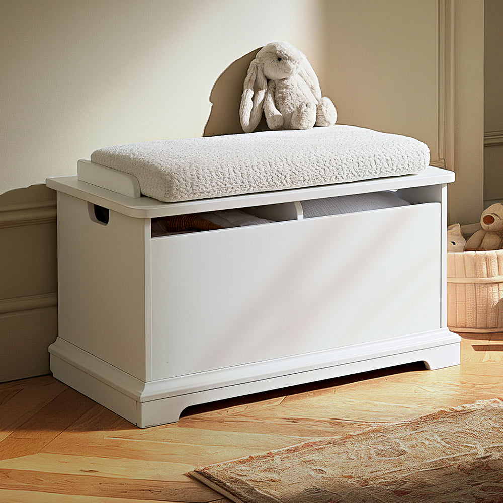 A white wooden toy storage bench with a cushioned seat, styled in a child's room with a plush bunny toy on top, a wooden floor, and a rug nearby.